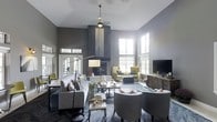 This single-floor space makes a strong architectural impression with its dramatically high ceilings, which lend an open, airy quality to the interior. The focal point of the main living area is a floor-to-ceiling fireplace clad in a striking chevron-patterned tile surround in muted gray tones, anchoring the room with both visual weight and warmth.

Large windows on multiple walls allow abundant natural light to filter throughout the space, complementing the neutral gray color palette that dominates the walls and soft furnishings. The layout is thoughtfully arranged with multiple seating zones, including plush gray sofas, tufted accent chairs, and mustard-yellow armchairs that introduce a pop of color against the otherwise subdued palette.

A media console with a flat-screen television is positioned along one wall, balancing the fireplace on the opposite side. French doors near the entrance provide a connection to the exterior, enhancing the sense of openness. Layered lighting, including a pendant fixture and floor lamps, adds depth and warmth. The overall ambiance is contemporary and refined, combining comfort with understated elegance.