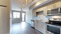 This single-floor unit showcases a contemporary open-concept layout that seamlessly connects the kitchen and living area. The kitchen is a standout feature, equipped with a full suite of stainless steel appliances, including a refrigerator, range, microwave, and dishwasher. Light wood-toned, flat-front cabinetry extends to the upper walls, maximizing storage while maintaining a clean, modern aesthetic. Speckled granite countertops complement the neutral color palette and provide ample workspace. Decorative amber glass pendant lights hang above, adding warmth to the space, while track lighting along the ceiling ensures functional illumination throughout the kitchen.

The living area flows naturally from the kitchen, featuring dark hardwood-style flooring that runs throughout the unit, creating visual continuity. A ceiling fan is centrally positioned, contributing to the room's comfort. A large window and sliding glass door provide abundant natural light and lead to a private balcony, offering outdoor views. The walls are finished in soft gray tones, lending a calm, neutral backdrop that complements the unit's modern design elements throughout.