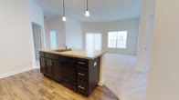Located at 100 Isabelle Isle in Dover, Delaware, this single-floor residence offers a thoughtfully designed open-concept layout that seamlessly connects the kitchen and living areas. The kitchen is anchored by a substantial dark-cabineted island topped with light granite countertops, providing ample workspace and storage through multiple drawers and cabinet doors. A built-in sink with a modern faucet and an integrated dishwasher add functional convenience. Pendant lighting hangs above the island, offering focused illumination with a clean, contemporary aesthetic. The kitchen transitions naturally into the living area, which is finished with plush carpeting and bathed in natural light from multiple windows dressed with vertical blinds. The contrast between the warm hardwood-style flooring in the kitchen and the soft carpet in the living space creates a clear yet cohesive delineation between the two zones. Neutral white walls throughout keep the interior feeling bright and open, while the high ceilings contribute to an airy, spacious atmosphere. The overall design emphasizes practicality and modern comfort, making this a well-appointed single-level home in a residential Dover neighborhood.