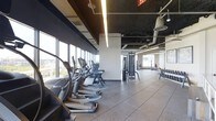 This fitness center offers a comprehensive workout environment spread across a single open floor. The space is defined by a wall of floor-to-ceiling windows along one side, flooding the interior with natural light and providing sweeping views of the surrounding cityscape. The exposed ceiling with visible ductwork and linear pendant lighting gives the facility a modern, industrial aesthetic.

The cardio zone lines the windowed wall and features a row of elliptical trainers and treadmills, allowing users to exercise while enjoying the outdoor views. Moving deeper into the space, a strength training area includes a cable and pulley machine system suitable for a variety of resistance exercises. A dedicated free weights section is visible toward the rear, equipped with a dumbbell rack offering a range of weight options.

The flooring throughout consists of durable rubber tiles, providing cushioning and stability appropriate for high-impact activity. The open layout ensures ample circulation space between equipment stations. The combination of natural light, contemporary finishes, and a well-organized equipment arrangement creates a functional and inviting environment suited for a full range of fitness routines.