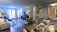 This single-floor residence is designed with an open-concept floor plan that seamlessly connects the living, dining, and kitchen areas, creating a cohesive and functional living environment. The living room is a welcoming space, anchored by rich hardwood flooring and a patterned area rug that adds texture and visual interest. Sheer curtains line the large windows, allowing generous natural light to fill the room while maintaining an airy, soft ambiance. Crown molding along the ceiling adds a refined architectural detail throughout.

The dining area sits adjacent to the living room, offering a natural flow for everyday living and entertaining. A wooden dining table with upholstered seating provides a practical yet stylish setting for meals.

The kitchen is accessible via a pass-through counter with a granite or stone-topped bar, offering both additional seating and an open sightline into the main living space. The kitchen features white cabinetry and a bright, well-lit workspace. Additional doors visible throughout suggest access to a bathroom, closet, or utility spaces, contributing to the overall practicality of the layout.

Neutral wall tones and consistent flooring materials unify the space, creating a calm, modern aesthetic that is both functional and inviting.