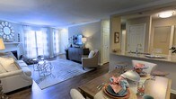 This single-floor residence is designed with an open-concept floor plan that seamlessly connects the living, dining, and kitchen areas, creating a cohesive and functional living environment. The living room is a welcoming space, anchored by rich hardwood flooring and a patterned area rug that adds texture and visual interest. Sheer curtains line the large windows, allowing generous natural light to fill the room while maintaining an airy, soft ambiance. Crown molding along the ceiling adds a refined architectural detail throughout.

The dining area sits adjacent to the living room, offering a natural flow for everyday living and entertaining. A wooden dining table with upholstered seating provides a practical yet stylish setting for meals.

The kitchen is accessible via a pass-through counter with a granite or stone-topped bar, offering both additional seating and an open sightline into the main living space. The kitchen features white cabinetry and a bright, well-lit workspace. Additional doors visible throughout suggest access to a bathroom, closet, or utility spaces, contributing to the overall practicality of the layout.

Neutral wall tones and consistent flooring materials unify the space, creating a calm, modern aesthetic that is both functional and inviting.