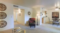 This single-floor space presents a well-organized, open layout that connects the living area to adjacent rooms with ease. The living room is carpeted and features neutral, warm-toned walls that create a bright and welcoming atmosphere. A decorative sunburst mirror serves as a focal point on the main wall, complemented by a classic armchair with an ottoman and a coordinating floor lamp, adding character to the space. A glass-topped coffee table anchors the seating area, enhancing the open feel of the room.

A wide, open doorway leads down a hallway lined with light hardwood-style flooring, providing a visual contrast to the carpeted living space and offering a sense of depth and connectivity throughout the unit. To the right, a partial wall partially encloses what appears to be a kitchen area, featuring a raised counter that provides a natural division while maintaining an open connection to the living space. Overhead ventilation registers are neatly integrated into the ceiling, contributing to the clean and functional design. Overall, the space balances practicality with a warm, lived-in aesthetic.