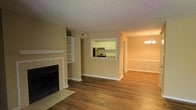 This single-floor residence offers a well-connected open-concept layout centered around a spacious living area. The living room is anchored by a classic fireplace with a white-trimmed mantle and tiled hearth, flanked by built-in shelving that provides both display and storage options. Rich hardwood flooring extends throughout, adding warmth and continuity to the space.

A pass-through window cut into the shared wall connects the living area directly to the kitchen, promoting an open, airy feel while maintaining a subtle separation between the two spaces. The kitchen features white cabinetry, granite-style countertops, and stainless steel appliances, including a refrigerator and range, making it both functional and visually clean.

Adjacent to the living area, a dedicated dining space is distinguished by an elegant chandelier, adding a refined touch to the overall aesthetic. Chair rail molding lines the walls, contributing an element of traditional architectural detail. Neutral beige walls and consistent white trim throughout the unit create a cohesive, inviting atmosphere well-suited to a variety of interior styles.