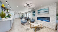 Located at 3221 W El Camino Dr in Phoenix, AZ, this single-floor professional office suite offers a thoughtfully designed workspace that balances modern style with functional efficiency. The welcoming reception and lounge area serves as the heart of the space, anchored by a striking stacked-stone accent wall housing a linear electric fireplace and framed coastal artwork. Sculptural ring pendant lights hang above comfortable seating arrangements, including blue accent chairs and a marble-top coffee table with gold accents, all set over a textured area rug and large-format tile flooring. Natural light filters in through multiple windows, brightening the open, airy atmosphere throughout. A sleek barn door with frosted glass panels adds a contemporary architectural touch, providing privacy between areas. The two private offices are well-appointed with large wood desks, dual-monitor setups, ample storage, and plantation shutters offering light control. A dedicated wet bar area features blue subway tile, open shelving, and a countertop sink — ideal for client hospitality. The space also includes a full bathroom and additional utility areas, making it a complete and polished professional environment suited for a variety of business uses.
