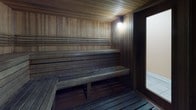 This single-room sauna is a well-appointed wellness space constructed entirely with natural wood, creating a warm and enveloping atmosphere. The walls, ceiling, and floor are all clad in horizontal wood planking, lending the interior a cohesive, traditional Scandinavian aesthetic. The wood tones range from lighter natural hues on the upper walls and ceiling to deeper, richly darkened tones on the lower benches and flooring, reflecting years of heat exposure.

The seating arrangement follows a classic tiered layout, with two levels of wide, slatted wooden benches running along the back and side walls. The upper bench offers a higher position for more intense heat, while the lower bench provides a cooler, more moderate experience. Both tiers comfortably accommodate multiple users simultaneously.

A traditional sauna heater is positioned near the entrance, serving as the functional centerpiece of the space. The entrance features a full-length glass-panel door framed in dark wood, allowing a visual connection to the tiled anteroom beyond while maintaining the thermal seal of the sauna interior. A wall-mounted light fixture provides soft, ambient illumination that complements the natural warmth of the wood. The overall design is straightforward, functional, and focused entirely on delivering an authentic and immersive sauna experience.
