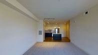 This single-floor space showcases an open-concept design that connects the living area to a well-appointed kitchen. The living area is finished with plush carpeting, providing a comfortable and inviting atmosphere, while the kitchen transitions to warm hardwood flooring. The kitchen is a standout feature, equipped with dark navy lower cabinetry contrasted by crisp white upper cabinets, creating a bold two-tone aesthetic. A central island with matching navy cabinetry serves as a functional focal point, ideal for casual dining or food preparation. Stainless steel appliances, including a full-sized refrigerator, complement the modern design. Overhead, track lighting and recessed ceiling lights illuminate the kitchen efficiently, while a decorative pendant light adds a refined touch above the island. A subtle tile backsplash adds texture behind the cooking area. The layout benefits from clean lines and a neutral color palette throughout, contributing to a bright, airy ambiance. The electrical panel is neatly integrated into the wall near the living space. Overall, the space offers a functional and stylish environment with a cohesive modern design.