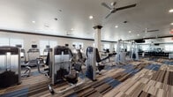 Located at 6600 McKinney Ranch Pkwy in McKinney, Texas, this single-floor fitness center is thoughtfully designed to support a comprehensive workout experience. The facility opens through a welcoming corridor lined with a striking multi-toned striped carpet in shades of blue, gray, and tan — a design element that carries throughout the entire space, adding visual energy underfoot.

The main workout floor is expansive and open, housing an extensive array of strength-training machines, free weights, and cable systems arranged for efficient circulation. Large wall mirrors amplify the sense of space, while multiple ceiling fans and recessed lighting maintain a comfortable, well-lit environment. Structural columns punctuate the floor plan, contributing to the architectural character of the space.

A dedicated cardio zone lines one side of the facility, featuring rows of treadmills, elliptical machines, and recumbent bikes positioned near windows that invite natural light. A separate room is equipped for spin classes, with stationary cycling bikes, stability balls, and a wall-mounted flat-screen TV for guided workouts. Another area accommodates rowing machines and specialty strength equipment for varied training needs.

The facility also includes conveniently placed water fountains, restroom access, and clearly marked exit signage, ensuring both comfort and safety throughout the space.