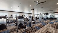 Located at 6600 McKinney Ranch Pkwy in McKinney, Texas, this single-floor fitness center is thoughtfully designed to support a comprehensive workout experience. The facility opens through a welcoming corridor lined with a striking multi-toned striped carpet in shades of blue, gray, and tan — a design element that carries throughout the entire space, adding visual energy underfoot.

The main workout floor is expansive and open, housing an extensive array of strength-training machines, free weights, and cable systems arranged for efficient circulation. Large wall mirrors amplify the sense of space, while multiple ceiling fans and recessed lighting maintain a comfortable, well-lit environment. Structural columns punctuate the floor plan, contributing to the architectural character of the space.

A dedicated cardio zone lines one side of the facility, featuring rows of treadmills, elliptical machines, and recumbent bikes positioned near windows that invite natural light. A separate room is equipped for spin classes, with stationary cycling bikes, stability balls, and a wall-mounted flat-screen TV for guided workouts. Another area accommodates rowing machines and specialty strength equipment for varied training needs.

The facility also includes conveniently placed water fountains, restroom access, and clearly marked exit signage, ensuring both comfort and safety throughout the space.