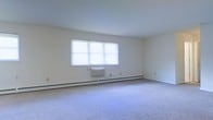 This single-floor space presents a clean, minimalist interior defined by its open layout and neutral color palette. The main room features wall-to-wall carpeting in a soft gray tone, providing a comfortable underfoot surface that contributes to the overall understated aesthetic. The walls are finished in a crisp off-white, enhancing the sense of brightness and openness throughout the space.

A large, multi-pane window serves as the focal point of the room, positioned centrally along one wall and fitted with white horizontal blinds. This window allows generous natural light to fill the interior, creating a well-lit and airy atmosphere. Beneath the window, a baseboard heating system runs the length of the wall, providing efficient climate control throughout the space. A wall-mounted air conditioning unit is also visible, ensuring year-round temperature management.

The room's smooth, flat ceiling and clean wall surfaces give the space a straightforward, functional character. Electrical outlets are evenly distributed along the walls, adding to the room's practical design. Overall, the space offers a neutral, move-in-ready environment suitable for a variety of uses.