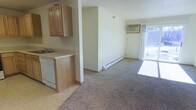 This single-floor space features an open-concept design that connects the kitchen and living area seamlessly. The kitchen is equipped with light wood cabinetry, a white dishwasher, a sink with a standard faucet, and light-toned countertops, all set on tile flooring. A partial wall separates the kitchen from the living area, providing a sense of definition without fully closing off the space. The living area transitions to wall-to-wall carpet and is heated by a baseboard heating unit running along the far wall. A sliding glass door floods the space with natural light and provides access to an outdoor area, offering views of the exterior surroundings. A framed piece of wall art adds a decorative touch near the living area. The textured ceiling is consistent throughout, and the neutral wall tones create a clean, understated ambiance. The layout is practical and functional, making efficient use of the available space across the single floor.