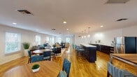 This single-floor community amenity space is thoughtfully designed to serve as a comfortable and functional gathering hub. The main area showcases warm hardwood flooring throughout, recessed lighting, and an abundance of natural light streaming in through multiple windows dressed with clean white blinds. The open-plan layout seamlessly connects a generously sized lounge area — furnished with plush seating in neutral tones accented by teal and navy blue — to a well-equipped kitchen and a variety of dining zones.

The kitchen features sleek dark cabinetry paired with crisp white countertops, a stainless steel refrigerator, dishwasher, and a large central island with bar seating illuminated by elegant pendant lights. Adjacent dining areas include both casual high-top tables and larger round tables with upholstered chairs, offering flexible seating arrangements.

A quieter lounge zone toward one end of the space provides a relaxing retreat, complete with armchairs and a coffee table bathed in natural light from surrounding windows. A dedicated laundry facility rounds out the amenities, housing multiple stacked and top-loading washers and dryers in a tidy, well-lit room with sage green walls and speckled flooring. A vending machine and additional storage areas are also accessible within the space, enhancing everyday convenience.