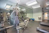 Located at 553 Settlers Landing Rd in Hampton, Virginia, this fitness center is a well-equipped, single-floor exercise facility designed to support a comprehensive workout routine. The space features neutral gray walls and durable rubber flooring throughout, providing a clean and functional environment suited for both cardio and strength training.

The cardio section is stocked with multiple treadmills, elliptical machines, and stationary bikes — including both upright and recumbent models — many of which are equipped with individual display screens for entertainment or performance tracking. A multi-station cable and weight machine anchors the strength-training area, offering users a range of resistance exercises in a compact footprint.

Full-length mirrors line one wall, visually expanding the space and allowing for form checks during workouts. Overhead, recessed fluorescent lighting panels provide bright, even illumination throughout the room. French-style doors with white blinds allow natural light to filter in while maintaining privacy, and contribute to an open, airy feel.

The layout is thoughtfully organized to maximize usable floor space, keeping equipment accessible and traffic flow clear. The overall ambiance is practical and professional, making this facility a convenient and well-appointed option for residents seeking a dedicated space for their fitness needs.