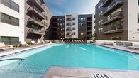 Located at 1701 Central Ave in the heart of Saint Petersburg, Florida, this outdoor amenity space is designed to deliver a resort-style experience within a contemporary multi-story residential community. The centerpiece is a generously sized swimming pool with crystal-clear blue water, surrounded by a sleek tiled deck lined with lounge chairs, providing ample space for sunbathing and relaxation under the Florida sun. Lush tropical plantings frame the pool area, complementing the warm climate and lending a vibrant, resort-inspired feel.

Adjacent to the pool is a thoughtfully designed outdoor lounge and entertainment area, anchored by a striking exposed brick accent wall mounted with a large flat-screen television. Comfortable seating, including modular sofas and colorful accent chairs, is arranged around a central table, creating an inviting space for socializing. Overhead pergola-style structures add architectural interest while providing partial shade.

A fully equipped outdoor kitchen with stainless steel grills, countertops, and a utility sink offers an ideal setup for alfresco dining and gatherings. The entire courtyard is paved with attractive stone pavers, tying the various amenity zones together cohesively. The surrounding multi-story building features contemporary dark and light paneling with private balconies, enclosing the space and contributing to a sense of community and privacy.