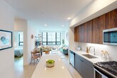 This thoughtfully designed single-floor residence offers a seamless open-concept layout that integrates the kitchen, dining, and living areas into one bright, cohesive space. The kitchen is a standout feature, showcasing rich walnut upper cabinetry paired with sleek gray lower cabinets, white quartz countertops, stainless steel appliances including a gas range, dishwasher, microwave, and refrigerator, and a central island that doubles as a casual gathering spot. The dining area flows naturally from the kitchen, accommodating a round table with modern wood chairs, while the living room beyond benefits from large, floor-to-ceiling windows with black frames that flood the space with natural light and provide urban views. Light hardwood flooring runs throughout, enhancing the airy, contemporary feel. The two bedrooms are well-proportioned, each featuring oversized windows, roller shades, and a clean, minimalist aesthetic. The bathroom is finished with large-format tile, a combination tub and shower, a gray vanity with dark countertop, and a wall-mounted mirror cabinet. Recessed lighting throughout the unit completes the polished, modern ambiance of this well-appointed residence.