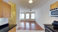 Located at The Corner Lofts, this single-floor unit blends classic character with modern conveniences. The kitchen is outfitted with stainless steel appliances, including a full-size refrigerator, electric range, and over-the-range microwave, complemented by warm-toned wood cabinetry and dark countertops. Light tile flooring in the kitchen transitions seamlessly into rich, wide-plank hardwood floors that extend through the living area, adding warmth and timeless appeal to the space. The living area is bright and open, benefiting from a trio of large windows that invite an abundance of natural light. Clean white walls enhance the airy atmosphere, while a ceiling fan with a light fixture provides both comfort and illumination. The generous proportions of the main living space offer flexible arrangement possibilities, making it well-suited for a variety of layouts. Crisp white baseboards and trim throughout add a polished finishing touch. Overall, the unit presents a welcoming and functional environment that combines the charm of original architectural details with practical, contemporary finishes.