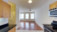 Located at The Corner Lofts, this single-floor unit blends classic character with modern conveniences. The kitchen is outfitted with stainless steel appliances, including a full-size refrigerator, electric range, and over-the-range microwave, complemented by warm-toned wood cabinetry and dark countertops. Light tile flooring in the kitchen transitions seamlessly into rich, wide-plank hardwood floors that extend through the living area, adding warmth and timeless appeal to the space. The living area is bright and open, benefiting from a trio of large windows that invite an abundance of natural light. Clean white walls enhance the airy atmosphere, while a ceiling fan with a light fixture provides both comfort and illumination. The generous proportions of the main living space offer flexible arrangement possibilities, making it well-suited for a variety of layouts. Crisp white baseboards and trim throughout add a polished finishing touch. Overall, the unit presents a welcoming and functional environment that combines the charm of original architectural details with practical, contemporary finishes.