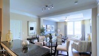 This single-floor residence showcases a thoughtfully designed open-concept layout that integrates the dining and living areas into one cohesive, flowing space. The dining area features a round glass-top table paired with upholstered cream chairs, anchored beneath a multi-head track lighting fixture that provides focused, ambient illumination. A dark granite countertop is visible in the foreground, hinting at a well-appointed kitchen nearby.

The transition into the living area is defined by a subtle architectural soffit, beyond which a tray ceiling adds dimension and visual interest. A ceiling fan with a light kit enhances comfort and airflow in the living space. Large windows dressed with layered curtains allow generous natural light to fill the room, creating a bright and airy atmosphere.

The living area is furnished with a neutral-toned sofa and complementary accent chairs, accessorized with decorative lamps and framed wall art. A console table along one wall is topped with a mirror and decorative accents, adding depth to the space. Lush indoor plants introduce a natural element throughout.

The overall color palette of soft beiges, warm grays, and whites creates a calm, sophisticated ambiance, making this space both visually appealing and functionally well-organized.