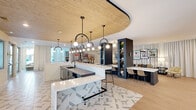 This thoughtfully designed single-floor amenity space offers a generous, open layout that seamlessly blends social and lounge-style living across several distinct zones. The centerpiece is a striking kitchen and lounge area anchored by dual islands with sleek white quartz countertops, dark cabinetry, and integrated appliances. A dramatic curved wood-paneled ceiling canopy overhead is punctuated by a series of arched pendant lights with globe bulbs, creating a warm, sculptural focal point. Adjacent dark navy built-in shelving units add architectural depth and display curated décor. The main living room stretches along a wall of floor-to-ceiling drapery, behind which a gallery-style arrangement of framed artwork adds vibrant color. A long communal work counter with bar seating runs parallel, ideal for collaborative use. Additional lounge nooks feature tall windows with courtyard and pool views, filling each seating area with natural light. Light hardwood flooring flows throughout, complemented by geometric-patterned tile in the kitchen zone. A secondary lounge area includes a wet bar with mosaic tile backsplash and a bold large-format art piece. The overall ambiance is contemporary and inviting, balancing stylish design with functional, multi-use comfort.
