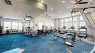 Located in Pflugerville, Texas, this well-appointed fitness center offers a comprehensive workout environment designed to support a wide range of exercise routines. The single-floor space is generously sized, providing ample room for members to move freely between equipment zones without feeling crowded.

The main workout area is anchored by vibrant blue and chevron-patterned carpet tiles that add a dynamic, energetic aesthetic to the space. Multiple ceiling fans are distributed throughout, ensuring consistent airflow and comfort during workouts. Recessed lighting, combined with an abundance of natural light streaming through large windows along multiple walls, creates a bright and inviting atmosphere.

The cardio section features a lineup of treadmills, ellipticals, stationary bikes, and stair climbers, all positioned to take advantage of the natural light. A dedicated spin area includes stationary cycling bikes paired with a wall-mounted flat-screen display running the Wellbeats virtual fitness platform, enabling guided on-demand classes. The strength training zone is equipped with cable machines, multi-function weight stations, benches, and a full dumbbell rack, complemented by floor-to-ceiling mirrors that visually expand the space. Additional accessories include stability balls, kettlebells, and a step platform. Built-in shelving and cabinetry near one wall provide organized storage and a polished finish to the overall design.