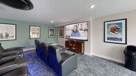 Located at 303 Dunbar St in Manchester, NH, this purpose-built home theater room delivers an immersive cinematic experience within a well-appointed single-floor layout. The room is designed exclusively for screening and entertainment, centered around a large wall-mounted display paired with a wooden media console that anchors the viewing wall. Two rows of tufted black leather recliner seats — each equipped with built-in cup holders and glowing blue LED underlighting — offer a true movie theater atmosphere and comfortable, stadium-style viewing for multiple guests. The flooring throughout is a durable, patterned commercial-grade carpet that complements the room's sleek, modern aesthetic. The walls are finished in a clean neutral palette with pops of sage green, and are adorned with framed movie posters that reinforce the cinematic theme. Recessed ceiling lighting provides even illumination, while a ceiling-mounted cassette HVAC unit ensures year-round climate control. A full-height glass partition wall along one side allows natural light to filter in from an adjacent corridor, maintaining an open and polished feel. Safety features including a fire extinguisher and emergency exit signage are also present. This is a thoughtfully designed entertainment space ideal for shared viewing experiences.