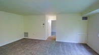 This single-story space presents a straightforward and functional layout, highlighted by wall-to-wall carpeting in a neutral gray tone that extends throughout the visible areas. The main living area is open and adequately sized, with smooth, light-colored walls that create a bright and clean atmosphere. A floor vent indicates a forced-air HVAC system integrated into the space.

A wide opening connects the main living area to an adjoining room, which appears to be an additional living space or bedroom, maintaining the same carpeted flooring and neutral color palette. The transition between spaces is seamless, contributing to an open and cohesive feel throughout.

White baseboards run along the perimeter of the rooms, adding a subtle but clean finish. The walls are painted in a soft, off-white tone, providing a versatile backdrop suitable for a variety of interior styles. A thermostat or utility panel is visible on one wall, indicating standard climate control functionality. The overall ambiance is understated and practical, with a blank-canvas quality that makes the space readily adaptable to the occupant's needs.