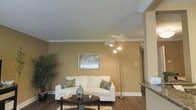 This single-story space presents a welcoming living area characterized by warm golden-toned walls and crisp white crown molding that frames the ceiling, adding a polished architectural detail throughout. The living room is centered around a comfortable white sofa accented with decorative paisley throw pillows, positioned atop a patterned area rug over dark hardwood flooring that adds richness and contrast to the space. A sleek multi-head arc floor lamp provides ambient and focused lighting, complementing the natural light that filters in through a window fitted with horizontal blinds. The textured white ceiling enhances the sense of openness in the room. Lush indoor greenery in the corner introduces a natural, refreshing element. An open passageway connects the living area to an adjacent space, which appears to feature dark countertops, suggesting a kitchen or bar area nearby. The overall ambiance is warm, casual, and functional, with a clean and well-maintained aesthetic that makes the space feel inviting and move-in ready.