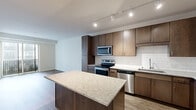 This thoughtfully designed 1-bedroom, 1-bathroom residence offers an efficient and modern layout across a single floor. The open-concept kitchen and living area serves as the heart of the home, featuring rich dark-toned cabinetry, a full suite of stainless steel appliances including a refrigerator, electric range, built-in microwave, and dishwasher. A white subway tile backsplash adds a clean, contemporary touch, while granite countertops extend across both the perimeter surfaces and a central island with built-in outlets — ideal for meal prep or casual dining. Track lighting illuminates the kitchen efficiently. The living area is finished with wood-look flooring and opens directly onto a private balcony through sliding glass doors, bringing in natural light and outdoor views. The bedroom is carpeted for comfort, features a large window for additional natural light, and includes access to a dedicated closet. The bathroom is finished to match the rest of the unit, with a dark wood vanity, granite countertop, undermount sink, and a large mirror. Throughout the space, neutral wall tones and consistent finishes create a cohesive, modern aesthetic.