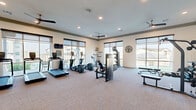 Located at 691 Union Chapel Rd in Cedar Creek, TX, this well-appointed fitness center offers a generously sized, single-floor exercise space designed to support a comprehensive workout experience. The room is flooded with natural light through large, floor-to-ceiling windows fitted with roller shades, providing views of the surrounding community and outdoor amenity areas, including a visible pool. The bright, neutral-toned interior features plush carpeted flooring, high ceilings with crown molding details, and multiple ceiling fans that maintain comfortable airflow throughout. Recessed lighting supplements the abundant natural light for evening use. The cardio zone is well-equipped with a row of treadmills, elliptical trainers, and a stationary bike, all positioned to face the expansive windows. A dedicated strength-training area includes cable machines, a leg press, adjustable weight benches, and a dumbbell rack with organized weight storage. Large wall-mounted mirrors span multiple walls, enhancing the sense of space and allowing users to monitor their form. Wall-mounted flat-screen televisions add an entertainment element to the cardio section. Decorative touches such as a wall clock and floating shelves contribute to the polished, community-oriented aesthetic of this functional and inviting fitness facility.