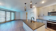 This well-appointed single-floor residence showcases a thoughtful open-concept layout that seamlessly connects the kitchen and living area. The kitchen is a standout feature, equipped with stainless steel appliances including a French door refrigerator, slide-in range, and over-the-range microwave. Gray shaker-style cabinetry with matte black hardware frames a striking herringbone tile backsplash, while a central island with a dark quartz countertop and undermount sink—topped by elegant geometric pendant lights—anchors the space. Light hardwood-style flooring flows throughout the main living areas, creating a warm and cohesive aesthetic. The open living area is bright and generously proportioned, with natural light filtering in through large windows dressed with blinds. The bedrooms are carpeted for comfort and feature ceiling fans, ample natural light, and access to walk-in closets with built-in shelving. The bathrooms are finished with light wood vanity cabinetry, dark-framed mirrors, quartz countertops, undermount sinks, matte black fixtures, and neutral tile flooring. One bathroom features a tiled shower surround, adding a refined touch. Throughout the space, clean white walls and modern hardware create a cohesive, contemporary atmosphere.