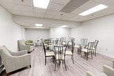 Located at 935 Dundas St E in Mississauga, Ontario, this single-floor commercial space offers a clean, functional, and welcoming environment suitable for a variety of uses. The interior is finished in a light, neutral palette throughout, with white walls and warm-toned laminate flooring that together create a bright and airy atmosphere.

The main open area serves as a generously sized lounge or gathering space, furnished with multiple round glass-top tables paired with elegant wrought-iron chairs and cream-cushioned seating. Comfortable upholstered armchairs and a small sofa are positioned along the perimeter, adding a relaxed, hospitality-oriented feel. A wall-mounted television provides an additional amenity for occupants.

A dedicated kitchenette area features white cabinetry with ample storage, a countertop with a sink, a microwave, and a stainless steel refrigerator, making the space well-equipped for light catering or refreshment needs. A large white service counter or reception island separates this functional zone from the main lounge area.

Throughout the space, a suspended drop ceiling with large recessed panel lighting ensures consistent, even illumination across all areas. The layout flows naturally from the main lounge to the kitchenette and ancillary spaces, with interior doors providing access to additional rooms. The overall design is practical yet polished, suited for professional, community, or hospitality-oriented purposes.