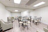 Located at 935 Dundas St E in Mississauga, Ontario, this single-floor commercial space offers a clean, functional, and welcoming environment suitable for a variety of uses. The interior is finished in a light, neutral palette throughout, with white walls and warm-toned laminate flooring that together create a bright and airy atmosphere.

The main open area serves as a generously sized lounge or gathering space, furnished with multiple round glass-top tables paired with elegant wrought-iron chairs and cream-cushioned seating. Comfortable upholstered armchairs and a small sofa are positioned along the perimeter, adding a relaxed, hospitality-oriented feel. A wall-mounted television provides an additional amenity for occupants.

A dedicated kitchenette area features white cabinetry with ample storage, a countertop with a sink, a microwave, and a stainless steel refrigerator, making the space well-equipped for light catering or refreshment needs. A large white service counter or reception island separates this functional zone from the main lounge area.

Throughout the space, a suspended drop ceiling with large recessed panel lighting ensures consistent, even illumination across all areas. The layout flows naturally from the main lounge to the kitchenette and ancillary spaces, with interior doors providing access to additional rooms. The overall design is practical yet polished, suited for professional, community, or hospitality-oriented purposes.