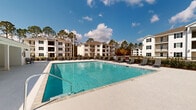 This outdoor pool facility in Biloxi, Mississippi offers a well-appointed recreational retreat designed for both relaxation and leisure. At the heart of the space is a generously sized swimming pool with clear, blue-tinted water, framed by a broad, light-colored concrete sundeck that provides ample room for lounging. Rows of outdoor chaise lounge chairs line the pool's perimeter, offering residents comfortable spots to unwind beneath the open sky.

A covered pavilion with crisp white columns and shiplap siding runs adjacent to the pool deck, providing a shaded gathering area that remains cool and inviting even on sunny days. The pavilion's open design allows for unobstructed views of the pool and the surrounding landscape of tall pines. The entire pool area is enclosed by a black metal fence, enhancing both safety and visual appeal.

A dedicated pool house structure sits nearby, clad in white shiplap siding and featuring large windows with shutters. It includes clearly marked, ADA-accessible men's and women's restrooms with glass-paned entry doors, ensuring convenience and accessibility for all users. The overall aesthetic is clean and contemporary, with a consistent white exterior palette that complements the natural, wooded backdrop of this Biloxi community.