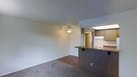 This single-floor space features an open-concept layout that seamlessly connects the living area to the kitchen. The living room is finished with wall-to-wall carpeting and transitions into hardwood-style flooring near the kitchen, creating a natural visual division between the two areas. A ceiling fan with a light fixture is centrally mounted, providing both air circulation and ambient lighting. The textured popcorn ceiling adds a classic touch throughout.

The kitchen is defined by a raised peninsula counter with dark wood panel detailing, offering a functional breakfast bar or prep space. Wood-toned cabinetry lines the kitchen walls, complemented by a built-in cooktop and an overhead range hood. A skylight above the kitchen brings in natural light, brightening the cooking area. A hallway visible in the background leads to additional rooms, with a spacious closet visible from the main living space. The neutral wall tones and understated finishes throughout give the space a clean, functional character well-suited for everyday living.