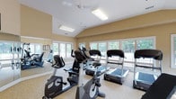 This fitness center is designed to provide a comprehensive workout environment within a bright, welcoming space. The room features vaulted ceilings that lend an open, airy feel, while warm beige walls and light-toned carpeted flooring create an inviting atmosphere. A ceiling fan and recessed flush-mount lighting fixtures ensure comfort and visibility throughout.

Large, multi-pane windows line multiple walls, flooding the space with abundant natural light and offering views of the surrounding greenery. Full-length mirrors are strategically placed along the walls, visually expanding the room and allowing users to monitor their form during exercise.

The cardio section is well-appointed with multiple treadmills, upright and recumbent stationary bikes, and elliptical machines from reputable brands including Spirit Fitness. A strength training station with a cable machine and a dedicated free weights area—featuring a tiered dumbbell rack—round out the equipment offerings for a full-body workout experience.

A wall-mounted flat-screen television, paired with a wall clock, adds convenience and entertainment during workouts. The space also includes a framed artwork piece and safety features such as clearly marked exit signage. Natural light, ample equipment variety, and a spacious layout make this fitness center a functional and comfortable facility for daily exercise.