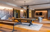 This expansive single-floor amenity lounge is designed with a contemporary aesthetic, combining open-plan living with distinct, purpose-driven zones. Wide-plank hardwood flooring flows throughout, unifying the space while bold navy-painted structural columns punctuate the layout, adding architectural character.

The games area serves as a central focal point, anchored by a pool table and shuffleboard table positioned atop a layered area rug beneath pendant lighting. Adjacent to this, a dedicated media lounge features a wall-mounted flat-screen television set against a dark textured accent wall, accompanied by comfortable sectional seating arranged for group viewing.

Multiple lounge seating areas are distributed throughout the space, furnished with generously sized sofas and accent chairs in neutral and earthy tones. Floor-to-ceiling windows and glass doors allow abundant natural light to fill the interior and provide direct access to an outdoor deck, where views of the surrounding urban skyline are visible.

A well-appointed wet bar area commands attention with its dramatic dark brick accent wall, veined marble countertops, a large waterfall-edge island, dark cabinetry with brass hardware, and vibrant contemporary artwork. Recessed and pendant lighting enhances the refined atmosphere throughout this sophisticated communal space.