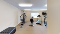This single-level fitness room offers a practical and functional space designed to support a variety of workout routines. The room features neutral beige walls, white trim, and carpeted flooring, creating a clean and comfortable environment for exercise. Overhead fluorescent lighting ensures the space is consistently well-lit, while windows along one wall allow natural light to supplement the artificial lighting and provide views of greenery outside.

The room is equipped with multiple treadmills, a stationary recumbent bike, and an upright cycle, offering ample options for cardiovascular training. A multi-station cable strength machine serves as the centerpiece for resistance-based workouts, complemented by a padded workout bench nearby. A wall-mounted flat-screen television is positioned for easy viewing during cardio sessions.

A large wall mirror spans a significant portion of one wall, visually expanding the space and allowing users to monitor their form during exercise. Additional features include a notice board, a yoga mat stored in the corner, and a dedicated entry door. The layout is organized and spacious, providing comfortable room to move between equipment.