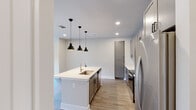 Located at 3710 Governors Dr SW in Huntsville, Alabama, this thoughtfully designed single-floor unit combines modern aesthetics with functional living spaces. The open-concept kitchen serves as the heart of the home, featuring a large center island with an undermount sink, crisp white quartz countertops, and a trio of matte black pendant lights overhead. Gray shaker-style cabinetry lines the walls alongside a classic white subway tile backsplash, complemented by stainless steel appliances including a refrigerator, range, and built-in microwave. Light-toned hardwood flooring flows seamlessly throughout the main living areas, enhancing the bright, airy atmosphere created by recessed lighting and natural light from windows and a glass-paneled exterior door. The living area offers a clean, open canvas with neutral walls and access to an outdoor balcony. Bedrooms are well-proportioned, featuring ceiling fans, ample natural light, and generous closet space with multiple built-in storage doors. Two bathrooms are finished with gray cabinetry, white quartz vanity tops, matte black hardware, large framed mirrors, and light tile flooring. One bathroom includes a glass-enclosed walk-in shower with built-in shelving, while the other features a full tub-shower combination. The overall design is cohesive, contemporary, and move-in ready.