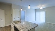 This single-floor space showcases an open-concept design that seamlessly connects the kitchen and living areas. The kitchen is anchored by a generously sized island topped with speckled granite countertops, featuring a double stainless steel undermount sink and a sleek pull-down faucet. A dishwasher is integrated beneath the island, and white cabinetry complements the neutral color palette throughout. Two cylindrical pendant lights hang above the island, providing focused task lighting while adding a modern aesthetic touch.

The open layout flows directly into the living area, which is finished with soft carpeting and painted in a consistent light gray tone that lends a calm, cohesive atmosphere. A ceiling fan with a built-in light fixture services this area, contributing to both comfort and functionality. A large window dressed with vertical blinds allows natural light to fill the space while offering privacy control.

A doorway visible from the kitchen leads to an additional room, suggesting a practical and well-organized floor plan. The overall ambiance is clean, contemporary, and move-in ready, with consistent finishes and neutral tones throughout.
