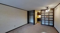 Located at 2350 Appleridge Dr in Columbus, Ohio, this single-floor residence offers a practical and functional layout designed to maximize its available space. The living area is finished with neutral-toned carpet and light-colored walls framed by dark baseboards and trim, creating a clean contrast throughout. A distinctive open shelving unit with a dark wood frame and frosted panel accents serves as a partial divider, subtly separating the living space from the adjacent areas while maintaining an open, connected feel. A ceiling fan is positioned overhead, contributing to the room's comfort and airflow. Built-in storage with dark cabinetry and a closet with bifold doors provides organized space along one wall, offering practical utility without compromising the open character of the room. The kitchen is visible beyond the divider, featuring dark upper cabinetry and under-cabinet lighting that casts a warm glow over the countertop workspace. The textured ceiling adds a layer of architectural detail consistent throughout the space. Overall, the residence presents a straightforward, functional interior with cohesive dark wood accents and neutral finishes that give it a grounded, utilitarian character.