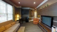 This single-floor space presents a thoughtfully designed interior that balances comfort with functionality. The main living area is anchored by a large flat-screen television mounted on a wood-paneled media wall, complemented by a sleek media console below. A plush sofa and an upholstered armchair in warm orange tones provide ample seating, while a patterned area rug adds texture and warmth to the carpeted floor.

Crown molding runs along the upper perimeter of the room, enhancing the refined aesthetic, while recessed ceiling lighting provides even, ambient illumination throughout the space. The walls are finished in warm earth tones, contributing to an inviting atmosphere.

A built-in kitchenette area is nestled along one wall, featuring dark cabinetry, a compact refrigerator, and a countertop workspace. Adjacent to this, a small bistro-style table with seating offers an intimate spot for individual use. Decorative artwork and framed pieces are displayed throughout, adding visual interest to the space.

A hallway visible in the background suggests additional areas beyond the main room, offering further utility and accessibility. The overall ambiance is cozy yet polished, making this space well-suited for relaxation or casual gatherings.