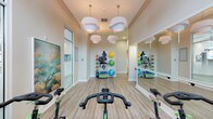 This single-floor yoga and fitness studio is thoughtfully designed to support a wide range of exercise disciplines in a clean, modern environment. The space features light wood-style flooring throughout, which lends warmth and a sense of openness to the room. Striking white pendant light clusters hang from the ceiling, providing bright, evenly distributed illumination that enhances the studio's welcoming ambiance.

A row of indoor cycling bikes is positioned along the mirrored wall, allowing riders to monitor their form during workouts. The full-length mirrors run the length of one wall and are complemented by a mounted ballet barre, making the space suitable for barre, dance, and stretching classes as well. Wall-mounted flat-screen TVs provide on-demand instruction and guided workout content directly in the cycling area.

A dedicated equipment zone houses an organized rack stocked with medicine balls, resistance bands, stability balls in multiple sizes, and additional accessories, offering versatility for strength and functional training sessions. Decorative large-scale artwork depicting serene cloudscapes and a motivational silhouette mural adds visual interest and an uplifting atmosphere. Floor-to-ceiling glass doors and transom windows invite natural light, keeping the studio feeling airy and energizing throughout the day.