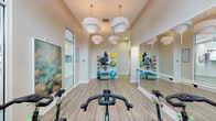 This single-floor yoga and fitness studio is thoughtfully designed to support a wide range of exercise disciplines in a clean, modern environment. The space features light wood-style flooring throughout, which lends warmth and a sense of openness to the room. Striking white pendant light clusters hang from the ceiling, providing bright, evenly distributed illumination that enhances the studio's welcoming ambiance.

A row of indoor cycling bikes is positioned along the mirrored wall, allowing riders to monitor their form during workouts. The full-length mirrors run the length of one wall and are complemented by a mounted ballet barre, making the space suitable for barre, dance, and stretching classes as well. Wall-mounted flat-screen TVs provide on-demand instruction and guided workout content directly in the cycling area.

A dedicated equipment zone houses an organized rack stocked with medicine balls, resistance bands, stability balls in multiple sizes, and additional accessories, offering versatility for strength and functional training sessions. Decorative large-scale artwork depicting serene cloudscapes and a motivational silhouette mural adds visual interest and an uplifting atmosphere. Floor-to-ceiling glass doors and transom windows invite natural light, keeping the studio feeling airy and energizing throughout the day.