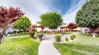 Located at 9924 Railroad Dr in El Paso, Texas, this residential community presents a thoughtfully designed outdoor environment that emphasizes comfort, greenery, and community living. The grounds are anchored by a central courtyard featuring a stone-edged circular landscaped focal point surrounded by mature deciduous trees, including striking red-leafed varieties that add seasonal color and visual interest throughout the property.

Wide, curved concrete walkways wind through the grounds, connecting residential buildings to communal gathering areas with ease of access. Red-painted pergolas provide shaded seating areas equipped with picnic tables, offering residents inviting spaces for outdoor relaxation and socializing. Covered walkways along building facades extend the usable outdoor space while providing shelter from the elements.

The landscaping throughout the community is meticulously maintained, blending lush green lawns with neatly trimmed shrubs, gravel ground cover, and ornamental plantings. Residential buildings feature light-colored vinyl siding with individual unit entrances, each serviced by dedicated HVAC units. The overall ambiance of the community is clean, organized, and welcoming, with abundant greenery softening the built environment and creating a pleasant, park-like atmosphere for residents to enjoy year-round.