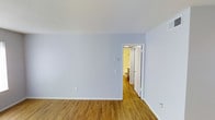 This single-floor space presents a straightforward and functional design characterized by clean, minimal finishes throughout. The main room features warm-toned hardwood-style flooring that extends seamlessly across the space, providing a cohesive and inviting foundation. The walls are painted in a soft, light blue-gray tone, contributing to a calm and neutral atmosphere that complements a variety of furnishing styles. White baseboards frame the perimeter of the room, adding a crisp contrast to the flooring and walls. The ceiling is smooth and white, enhancing the sense of openness within the room. A doorway with a white-framed door leads directly into an adjoining bathroom, which is visible from the main room and features warm yellow-toned walls, a toilet, and overhead lighting. The layout is practical, with the bathroom conveniently accessible from the primary living area. Electrical outlets and a thermostat panel are positioned along the walls, reflecting thoughtful placement for everyday functionality. Overall, the space offers a clean, neutral palette that is well-suited for versatile use.