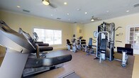 Located at 1000 Fawn Drive in Williamstown, NJ, this fitness center offers a well-equipped, single-floor exercise space designed to support a range of workout routines. The room features warm yellow walls accented by crisp crown molding, recessed lighting, and ceiling fans that maintain a comfortable environment during workouts. Natural light filters in through a large window, offering views of the surrounding community and keeping the space feeling open and inviting. The flooring combines durable carpet and rubberized mat sections, providing appropriate surfaces for both machine-based and functional training. The cardio area is outfitted with multiple recumbent bikes, an upright cycle, and treadmill-style equipment, arranged to allow ample circulation between machines. On the strength side, a multi-station cable and weight machine provides users with a comprehensive range of resistance training options. A mounted display screen is also present, offering instructional or entertainment support during exercise sessions. The layout is thoughtfully organized to maximize usable floor space while maintaining an uncluttered, accessible atmosphere suitable for residents of all fitness levels.