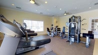 Located at 1000 Fawn Drive in Williamstown, NJ, this fitness center offers a well-equipped, single-floor exercise space designed to support a range of workout routines. The room features warm yellow walls accented by crisp crown molding, recessed lighting, and ceiling fans that maintain a comfortable environment during workouts. Natural light filters in through a large window, offering views of the surrounding community and keeping the space feeling open and inviting. The flooring combines durable carpet and rubberized mat sections, providing appropriate surfaces for both machine-based and functional training. The cardio area is outfitted with multiple recumbent bikes, an upright cycle, and treadmill-style equipment, arranged to allow ample circulation between machines. On the strength side, a multi-station cable and weight machine provides users with a comprehensive range of resistance training options. A mounted display screen is also present, offering instructional or entertainment support during exercise sessions. The layout is thoughtfully organized to maximize usable floor space while maintaining an uncluttered, accessible atmosphere suitable for residents of all fitness levels.