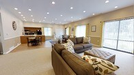 This single-floor space offers an open-concept layout that seamlessly connects a relaxed lounge area, a dining space, and a functional kitchenette. The lounge is furnished with plush, earth-toned sofas accented with botanical-patterned throw pillows, arranged over a neutral area rug to create a warm and inviting gathering area. An upholstered ottoman serves as a central accent piece, adding both comfort and style.

The dining area is positioned adjacent to the lounge and features a sturdy table with chairs, ideal for shared meals or casual meetings. Just beyond, the kitchenette is equipped with cabinetry, countertop appliances, and a refrigerator, providing practical amenities for residents or guests.

Throughout the space, large windows dressed with cheerful yellow-accented curtains allow natural light to filter in, complementing the warm beige wall tones and light hardwood-style flooring. An abundance of recessed ceiling lights ensures the space remains bright and well-lit at all hours. The overall ambiance is clean, functional, and welcoming — reflecting a thoughtfully designed communal environment suited for socializing, dining, and everyday gathering.