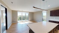 This well-appointed single-floor residence offers a thoughtfully designed open-concept layout that seamlessly connects the kitchen and living areas. The kitchen is a standout feature, showcasing rich dark-wood cabinetry, crisp white countertops, a large center island with pendant lighting, and a full suite of stainless steel appliances including a refrigerator, range, microwave, and dishwasher. Light wood flooring flows throughout the main living areas, complementing the neutral, warm-toned walls and creating a cohesive, inviting atmosphere.

The spacious living area is bathed in natural light, courtesy of large sliding glass doors that open onto a private balcony. The balcony offers sweeping views of the surrounding treetops and skyline, providing a serene outdoor retreat. Two generously sized bedrooms feature plush carpeting and large windows that frame elevated views of the landscape, ensuring brightness throughout the day.

The unit includes two bathrooms: one featuring a soaking tub and shower combination with dark wood vanity cabinetry, and a second with a walk-in glass-enclosed shower. Both bathrooms are finished with light wood flooring and warm neutral tones, maintaining the consistent, modern aesthetic found throughout the entire residence.