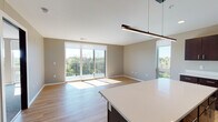 This well-appointed single-floor residence offers a thoughtfully designed open-concept layout that seamlessly connects the kitchen and living areas. The kitchen is a standout feature, showcasing rich dark-wood cabinetry, crisp white countertops, a large center island with pendant lighting, and a full suite of stainless steel appliances including a refrigerator, range, microwave, and dishwasher. Light wood flooring flows throughout the main living areas, complementing the neutral, warm-toned walls and creating a cohesive, inviting atmosphere.

The spacious living area is bathed in natural light, courtesy of large sliding glass doors that open onto a private balcony. The balcony offers sweeping views of the surrounding treetops and skyline, providing a serene outdoor retreat. Two generously sized bedrooms feature plush carpeting and large windows that frame elevated views of the landscape, ensuring brightness throughout the day.

The unit includes two bathrooms: one featuring a soaking tub and shower combination with dark wood vanity cabinetry, and a second with a walk-in glass-enclosed shower. Both bathrooms are finished with light wood flooring and warm neutral tones, maintaining the consistent, modern aesthetic found throughout the entire residence.